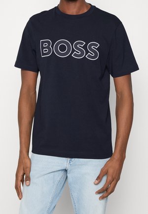 Mann trägt ein marineblaues T-Shirt mit weiß umrandetem "BOSS"-Schriftzug, kombiniert mit hellblauen Jeans, vor einem schlichten weißen Hintergrund.