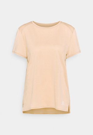 Beżowy t-shirt z krótkim rękawem i okrągłym dekoltem, z małym białym logo blisko dolnego brzegu, na jednolitym jasnym tle.