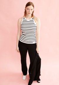 Top senza maniche a righe bianche e nere con texture a coste, abbinato a pantaloni neri aderenti. La modella tiene una giacca nera e indossa scarpe bianche.