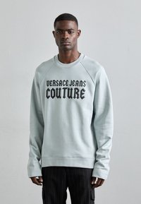 Světle modrý sweatshirt s kulatým výstřihem a raglánovými rukávy. Na hrudi je černý nápis "VERSACE JEANS COUTURE".