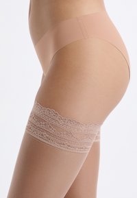 Nude pończochy do ud z gładkim, przezroczystym materiałem i ozdobnym koronkowym paskiem na górze z falistym brzegiem.