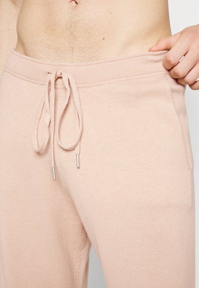 Barriga à mostra com pelos corporais claros acima de calças de moletom rosa pálido com cordão, seguradas por uma mão na cintura.