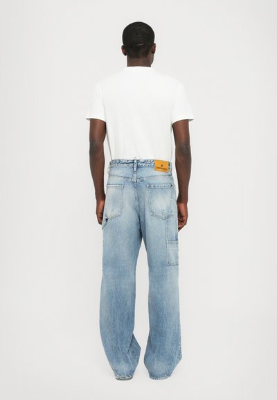 Ljusblå, vida denimjeans med förstärkt bakficka och en tan läderpatch i midjan. Slät textur, avslappnad design.