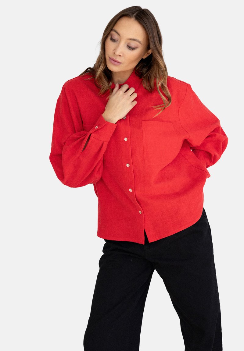 Czerwona koszula z długim rękawem, z pojedynczą kieszenią na piersi, o luźnym kroju. Materiał wydaje się mieć fakturę, stylizowana z czarnymi spodniami.