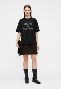 Γυναίκα που φοράει μαύρο μεγάλο μπλουζάκι με το κείμενο "ZADIG ET VOLTAIRE", φούστα σκούρου καφέ, μαύρες μπότες μέχρι τη μέση της γάμπας, κρατώντας μαύρο τσαντάκι με φούντες.