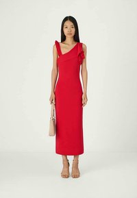 Donna in un vestito midi rosso a spalla scoperta con dettagli a volant, con una borsa beige in mano e sandali col tacco nudi.