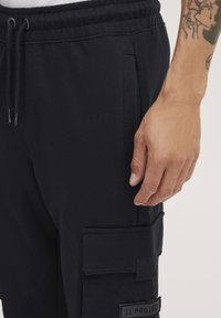 Zwarte sweats met elastische tailleband, trekkoord, zijzakken en een patchzak op de dij met een merklabel.