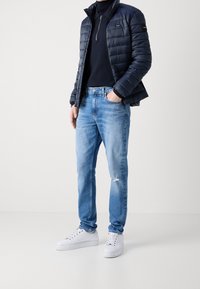 Vadderad marinblå jacka med dragkedja, svart polo, ljusblå slitna jeans och vita sneakers. Slim fit-design med en avslappnad stil.