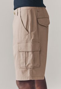 Shorts cargo beiges en coton, dotés de multiples poches, d'une fermeture par bouton et de détails de coutures visibles. Longueur mi-cuisse.