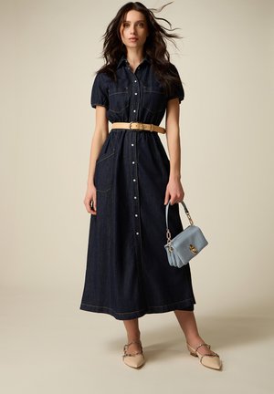 Kobieta w ciemnej dżinsowej sukience midi na guziki z krótkimi rękawami i brązowym paskiem, trzymająca jasnoniebieską torebkę, stojąca na beżowym tle.