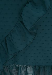 TÜRKISFARBENES TEXTIL MIT STRUKTURIERTER OBERFLÄCHE, DAS ERHOBENE PUNKTE AUFWEIST, MIT EINFALTEN AN DER KANTE UND EINEM FALTENBASIS, WAS EINE SCHICKE LAYERED OPTIK ERZEUGT.