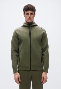 Sudadera con capucha y cremallera en color verde oliva, confeccionada en punto texturizado. Cuenta con cremallera negra y bolsillos laterales, con detalles acanalados en los hombros y las mangas.