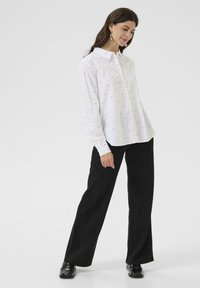Chemise blanche à boutons avec de petits motifs bleus, à manches longues et à col classique, associée à un pantalon noir à larges jambes et des chaussures noires.
