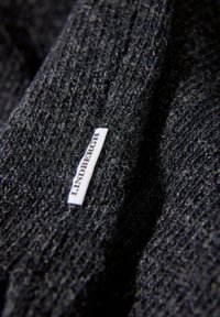 Svart strukturerad tyg med grov stickning, med en vit etikett där det står "LINDBERGH" i svart text sytt på sidan.