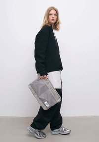 Sort kordfløjlskjorte over en hvid skjorte, parret med sorte vide bukser. Holder en grå mesh lynlås pung. Grå sneakers med accenter.