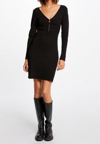 Černé žebrované šaty s dlouhým rukávem a výstřihem do V, s centrálním zipem, přiléhavý střih do poloviny stehna, doplněné černými kozačkami ke kolenům.