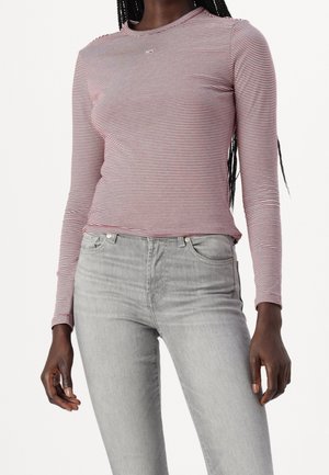 Frau trägt ein eng anliegendes, langärmeliges, rosa-weiß gestreiftes Hemd kombiniert mit hellgrauen Jeans vor einem einfarbigen Hintergrund.