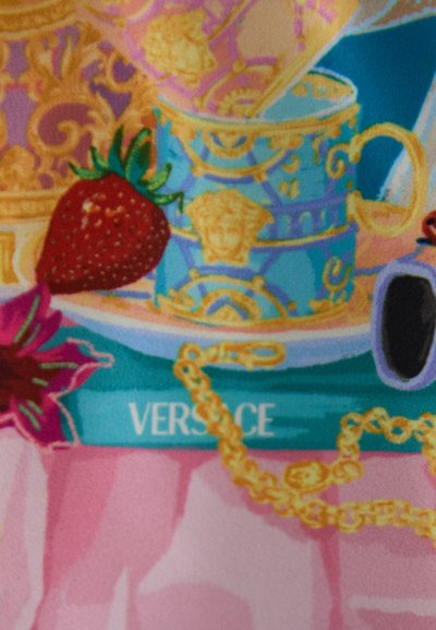 Versace mug in blue and gold with ornate designs, a red strawberry, floral accents, and a gold chain against a pink background.