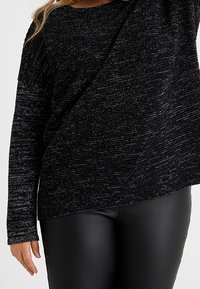 Žena v černém texturovaném svetr s dlouhými rukávy a černých koženkových kalhotách, snímek od ramen po boky.