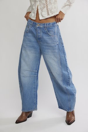 Naine, kellel on seljas laia säärega sinised teksapüksid, pruunid terava ninaga pahkluu saabastega ja valge pitsist pikkade varrukatega lühike top.