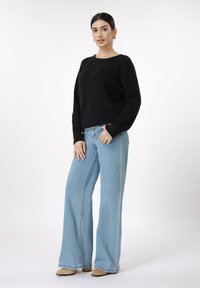 Μαύρο πλεκτό πουλόβερ με ριμπ υφή, στρογγυλή λαιμόκοψη και άνετη εφαρμογή, συνδυασμένο με ανοιχτό μπλε τζιν παντελόνι με φαρδιά πόδια και μπεζ παπούτσια.