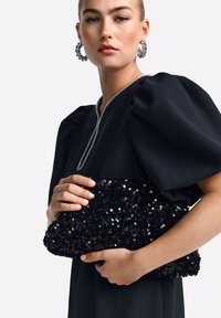 Fekete flitteres kézitáska, amelyet kezében tartanak, puha textúrával és kerek formával. A modellen fekete ruhát visel, amelynek puffos ujja van.