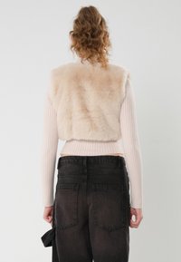 Vesta din blană sintetică în bej deschis, bluză cu mâneci lungi ribbed în roz deschis, combinată cu blugi largi decolorati. Design texturat, scurt.
