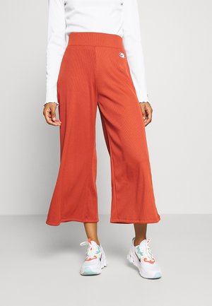 Rostfarbene Hosen mit weitem Bein und Rippenstruktur, hoher Taille und einem Logopatch an der Seite, kombiniert mit weißen Sneakers.