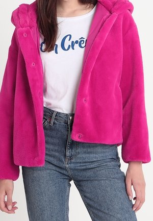 Femme portant une veste en peluche rose vif sur un t-shirt blanc imprimé et un jean bleu taille haute, debout, bras détendus.