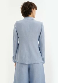 Hellblauer, taillierter Blazer mit langen Ärmeln, spitzen Revers und geknöpften Manschetten. Strukturierter Stoff; schmale Passform mit minimalen Details.