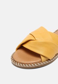 Sandale mule en cuir jaune avec une large bride torsadée, une semelle intérieure camel rembourrée et une bordure cousue sur une semelle plate, sur fond blanc.