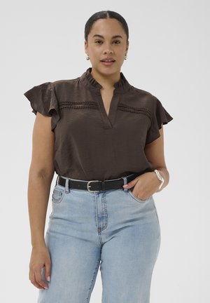 Femme portant un chemisier à volants manches courtes marron foncé, rentré dans un jean bleu clair avec une ceinture noire, debout avec une main dans la poche.