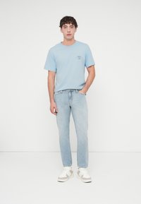 Ανοιχτό μπλε κοντομάνικο μπλουζάκι με γραφικό στην πρόσοψη, συνδυασμένο με ανοιχτό μπλε τζιν και λευκά αθλητικά παπούτσια, που στέκεται μπροστά σε απλό φόντο.