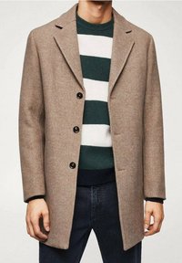 Beżowy płaszcz w jodełkę z klapami z wycięciem, czarnymi guzikami i dopasowanym fasonem, noszony na zielono-białym paskowanym swetrze.