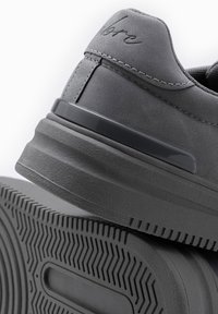 Zapatilla gris con parte superior sintética lisa, acentos cosidos y una suela de goma texturizada con un patrón de banda de rodadura en zigzag.