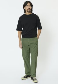 Camiseta negra de manga corta, pantalones plisados verde oliva y zapatillas negras con suelas de goma blancas. Corte casual, diseño minimalista.