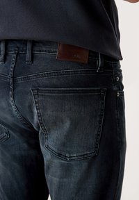Jeans de mezclilla de un azul oscuro con una textura suave, que presentan un parche de cuero con el logotipo, bolsillos traseros y sutiles detalles de desvanecimiento.