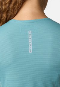 Una maglia sportiva turchese con un motivo a griglia testurizzato, dotata di un'etichetta bianca "SWEDMOUNT" sul retro vicino al colletto.