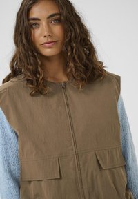 Barna mellény magas gallérral, két elülső zsebbel; világoskék kötött pulóverrel, amely texturált mintával rendelkezik.