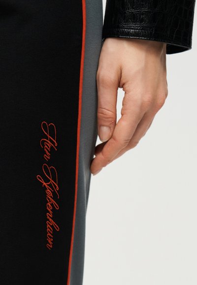 Χέρι δίπλα σε μαύρο και γκρι παντελόνι με κόκκινη περιμετρική λεπτομέρεια και το κέντημα "Han Kjøbenhavn" σε κόκκινη γραφική γραμματοσειρά.