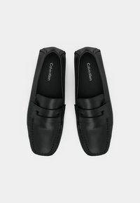 Μαύρα δερμάτινα παπούτσια με γλίστρημα, λεία υφή, τετράγωνη μύτη και λεπτομέρεια με λουράκι μπροστά. Εσωτερική επένδυση με ορατό το όνομα της επωνυμίας.