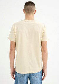 Camiseta beige de manga corta, hecha de una tela suave, con un dobladillo redondeado y detalles de costura sutiles, combinada con jeans de color azul claro.