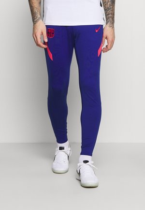 Mann trägt blaue Nike FC Barcelona Trainingshose und weiße Sneakers, steht vor einem schlichten hellen Hintergrund.