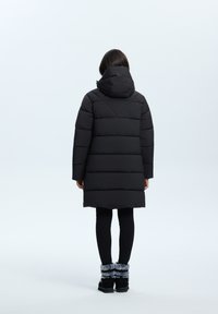 Zwarte pufferjas met capuchon, voorzien van horizontale gestikte secties, knielange stijl en een gladde, glanzende afwerking. Zwarte legging en laarzen.