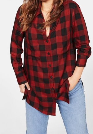 Kvinde iført en rød og sort ternet langærmet skjorte med sorte knapper og lyseblå jeans, står med den ene hånd i lommen.
