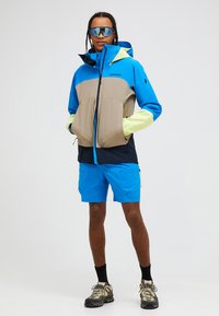 Bunte Sportjacke mit blau-beigem Design, langen Ärmeln und Kapuze. Kombiniert mit blauen Shorts und Outdoor-Schuhwerk.