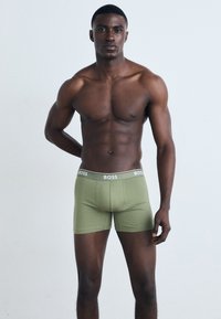 Grüne Baumwoll-Boxershorts mit einer enganliegendem Design, ausgestattet mit einem weißen Logo-Bund. Das Modell steht selbstbewusst mit entspannten Armen.