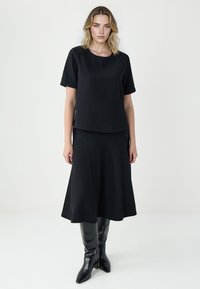 Kobieta stoi, ubrana w czarną bluzkę z krótkim rękawem, czarną spódnicę midi oraz czarne wysokie botki na obcasie, na jednolitym jasnym tle.