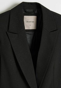 Czarna marynarka o delikatnej teksturze i subtelnym wzorze. Posiada klapowany kołnierz oraz widoczną metkę w środku z nazwą marki i rozmiarem.