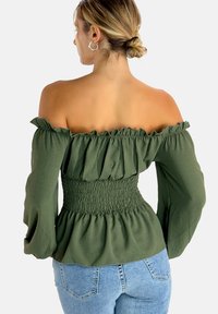 Off-the-shoulder olive green top with ruffled edges and a smocked waist. Long sleeves and a fitted silhouette. Textured fabric. Casual design.